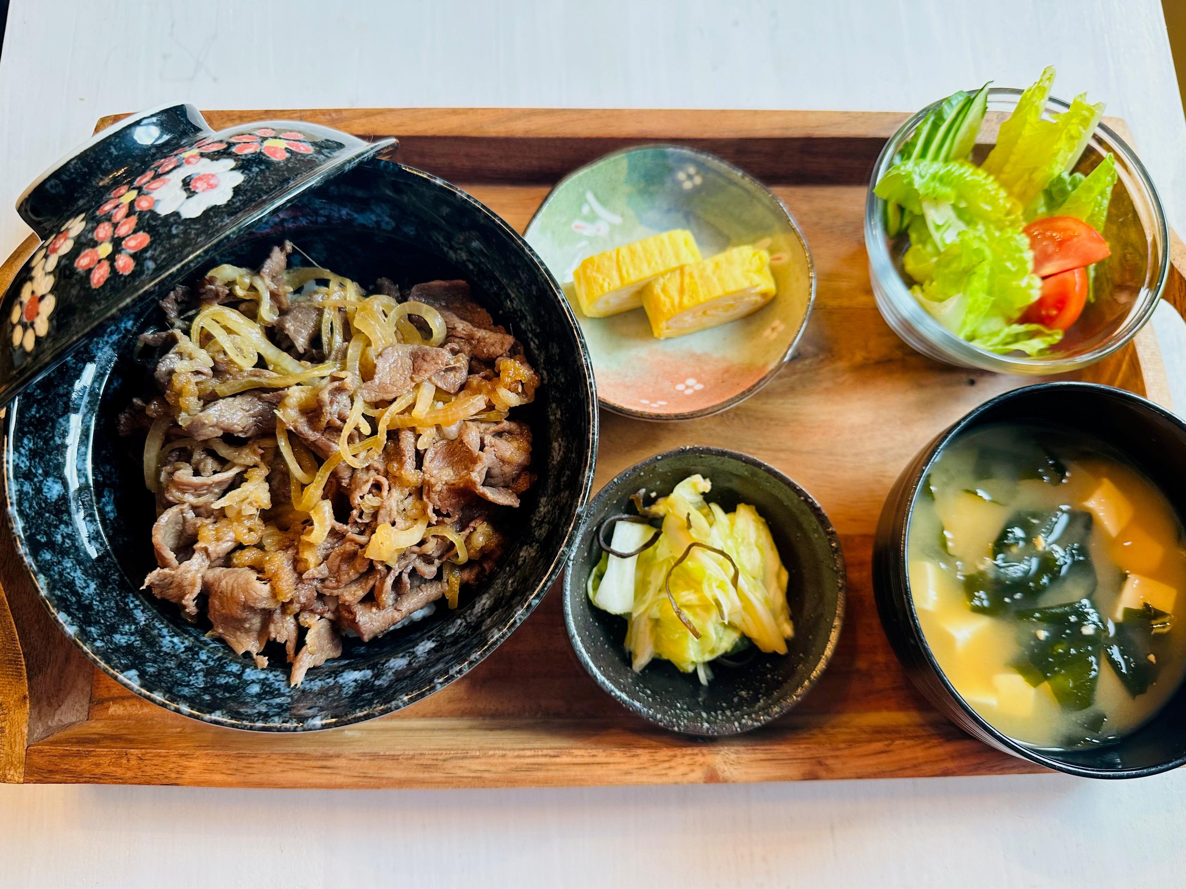 A Japanese set meal on a wooden tray: beef rice bowl, miso soup, rolled omelette, pickles, and salad